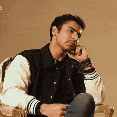 a man wearing a black and white varsity jacket sits in a chair with his hand on his chin