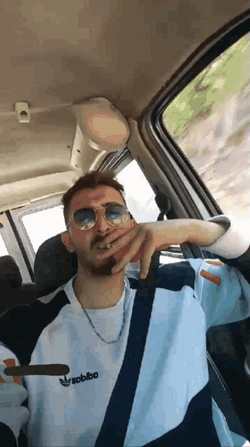 a man wearing sunglasses and an adidas shirt is smoking a cigarette while sitting in a car