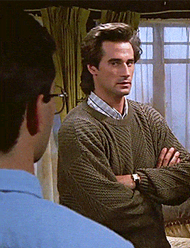 a man in a sweater stands with his arms crossed in front of another man in a blue shirt