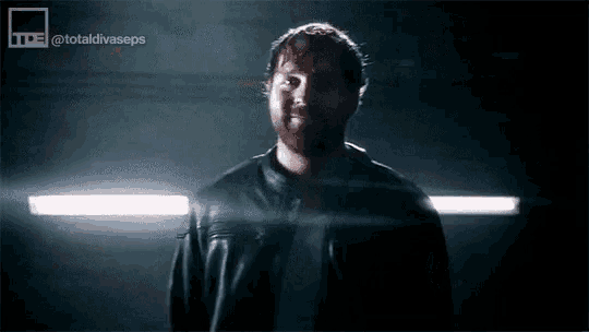 a man in a black leather jacket is standing in a dark room with lights behind him .