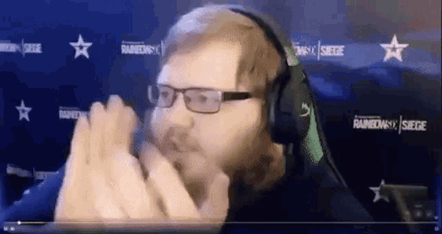 a man wearing glasses and headphones is giving a high five in front of a rainbow six siege sign .