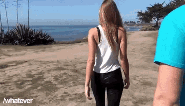 a woman in a white tank top and black pants is walking on a beach with a man in a blue shirt ..