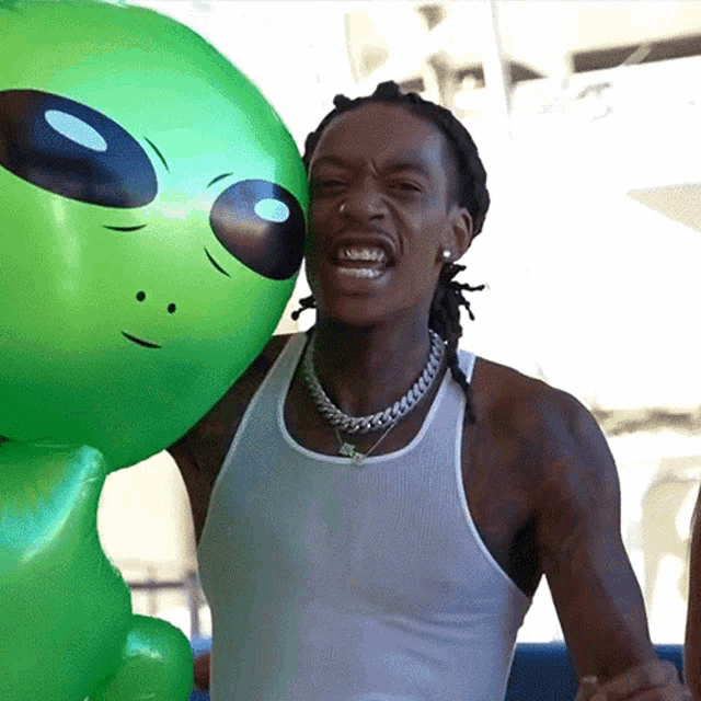 a man in a white tank top holds a green inflatable alien