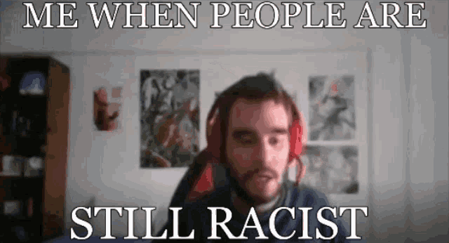 a man wearing headphones is sitting in front of a wall with posters on it and says me when people are still racist