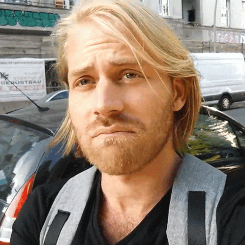 a man with long blonde hair and a beard looks at the camera in front of a sign that says gerustbau