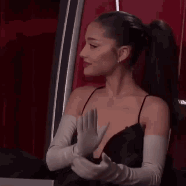 ariana grande is wearing a black dress and white gloves and sitting in a chair .