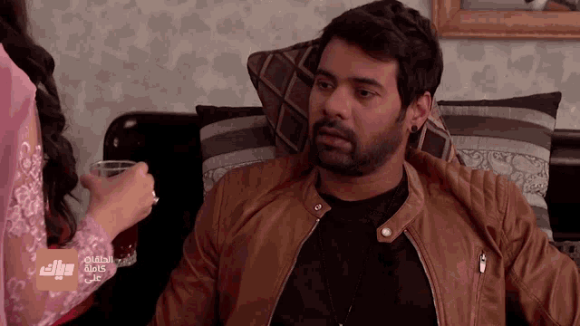 a man in a brown leather jacket sits on a couch with a woman holding a glass in front of him