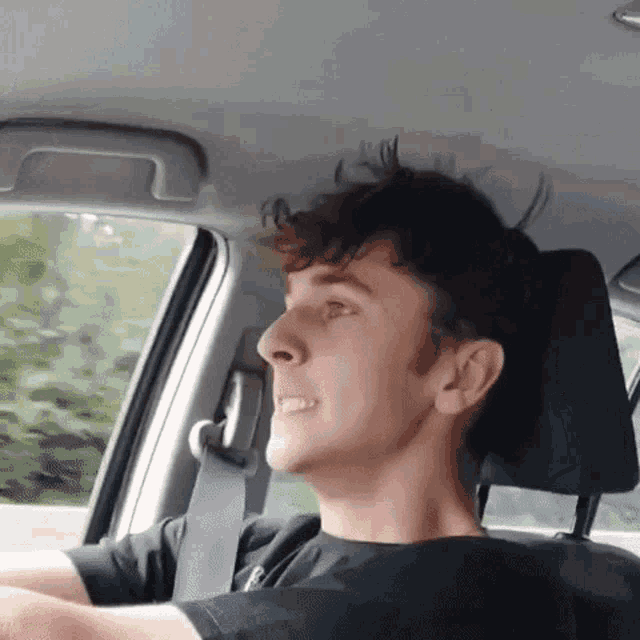a young man is sitting in the driver 's seat of a car with his hair blowing in the wind .