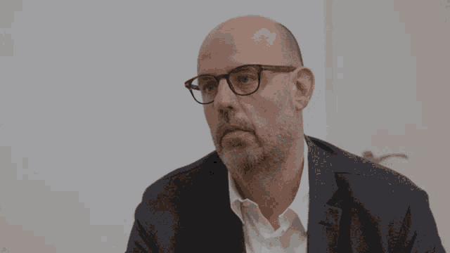 a bald man wearing glasses and a suit looks to the side