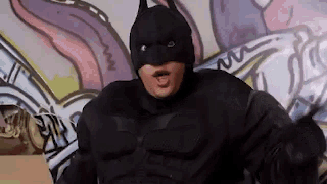 a man in a batman costume is making a funny face in front of a graffiti wall .