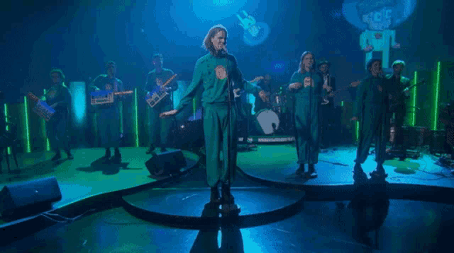 a group of people in green jumpsuits are playing instruments and singing on a stage