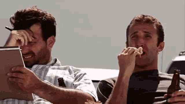 two men are sitting next to each other and one is eating a hot dog
