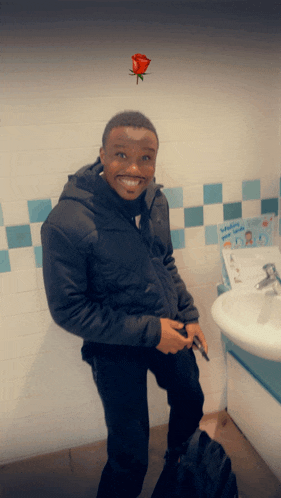 a man in a black jacket stands in front of a bathroom sink with a red rose on the wall above him