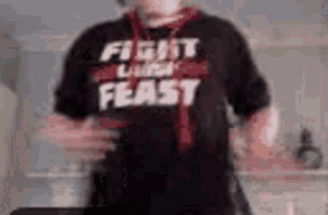 a man wearing a black shirt that says fight like a feast is standing in a kitchen .