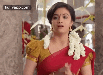 a woman in a red saree with a garland around her neck is standing in front of a wall .