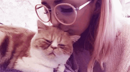 a woman with pink hair and glasses is kissing a cat on the nose .