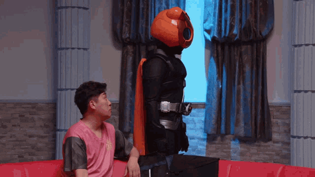 a man in a pink shirt sits on a couch next to a man in an orange costume