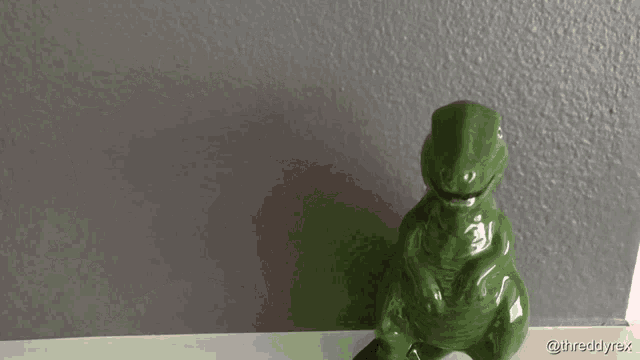 a green t-rex figurine sits on a shelf next to a grey wall