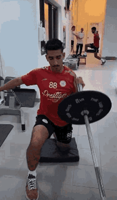 a man in a red shirt with the number 88 on it squats with a barbell