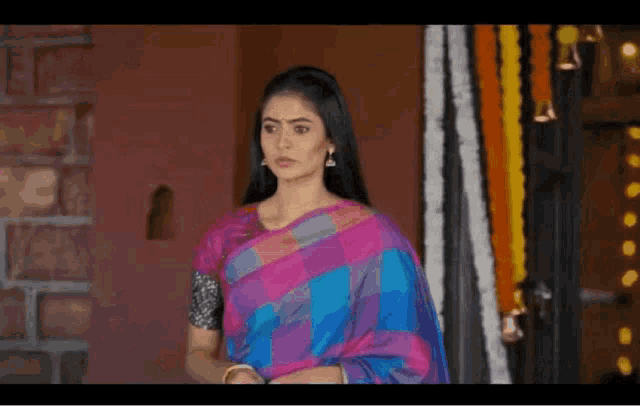 a woman in a pink and blue saree is standing in front of a brick wall .