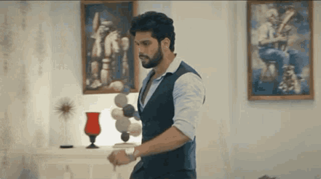 a man in a vest and white shirt is standing in a living room with paintings on the wall .