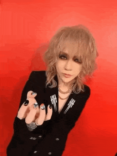 a woman with blonde hair and black nails is wearing a black jacket and making a hand gesture .