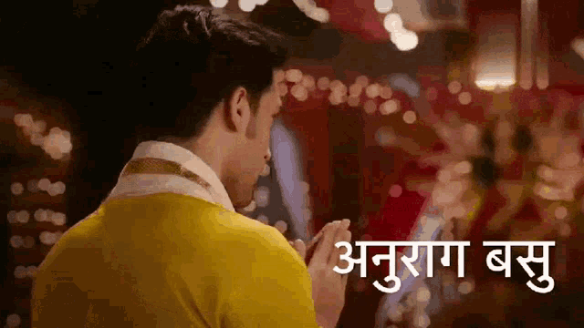 a man in a yellow shirt is praying in front of a blurred background and the words anurag basu are visible