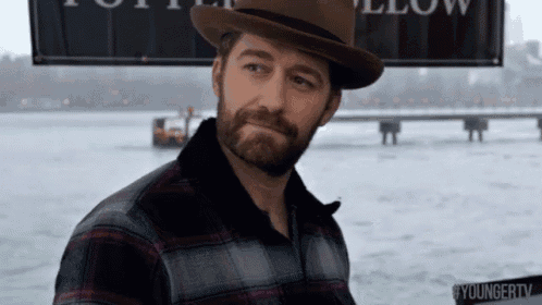 a man with a beard wearing a hat and a plaid shirt is standing in front of a body of water .
