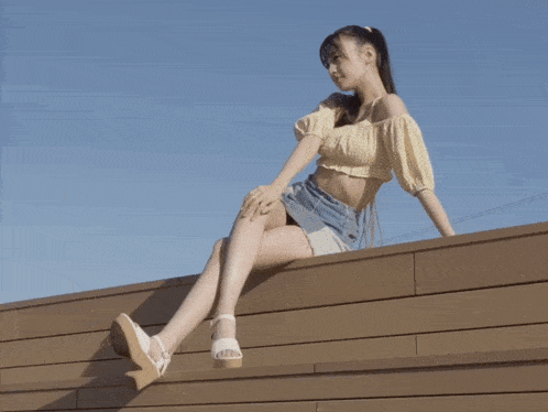 a woman in a yellow crop top and denim skirt sits on a wooden ledge