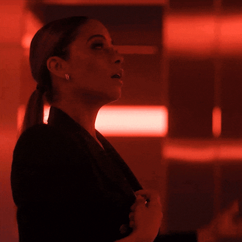 a woman in a black jacket stands in a dark room with a red light behind her