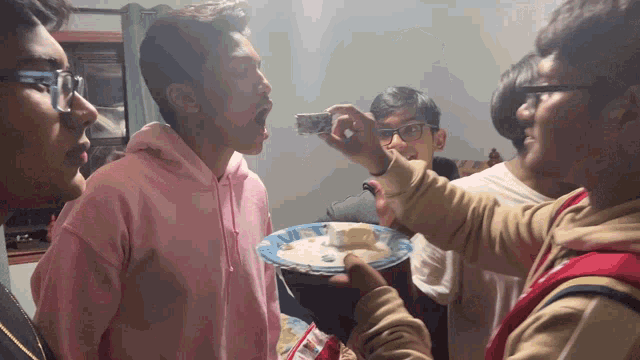 a man in a pink hoodie is being fed a piece of cake by another man in glasses