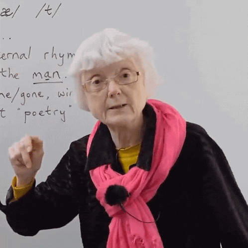 an elderly woman wearing glasses and a pink scarf is pointing at a white board with the word poetry written on it