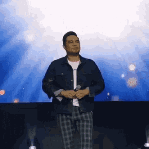 a man in a denim jacket and plaid pants is standing on a stage with a microphone .