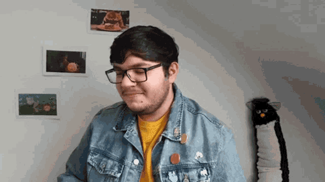a man wearing glasses and a denim jacket stands in front of a wall with pictures on it .