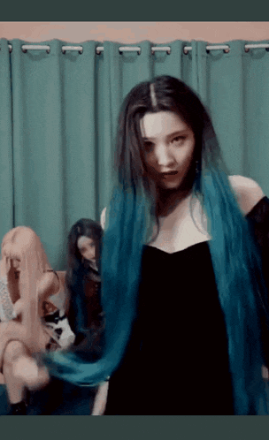 a woman with long blue hair is wearing a black dress and standing in front of a green curtain .