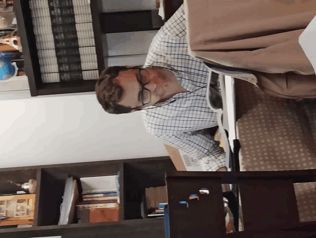 a man wearing glasses sits at a desk in front of a bookshelf with a few books on it
