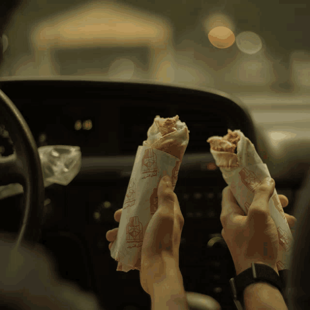 a person in a car is holding two sandwiches wrapped in paper that says ' arabic ' on it