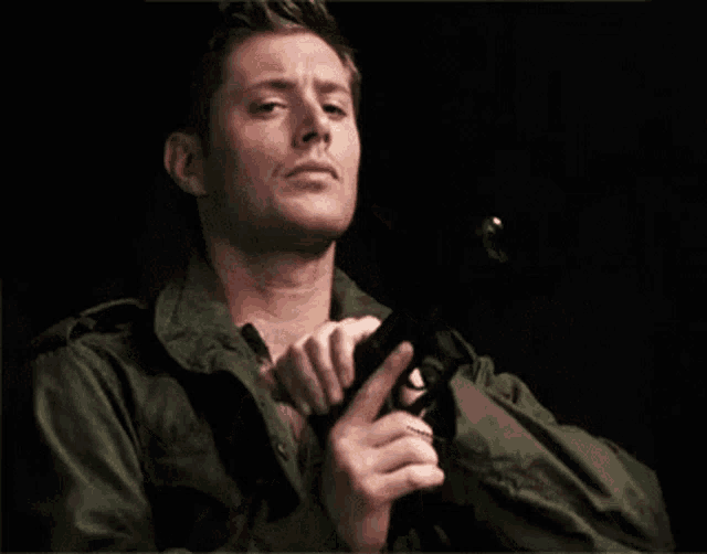 a man in a green jacket is holding a gun in his hand in a dark room .