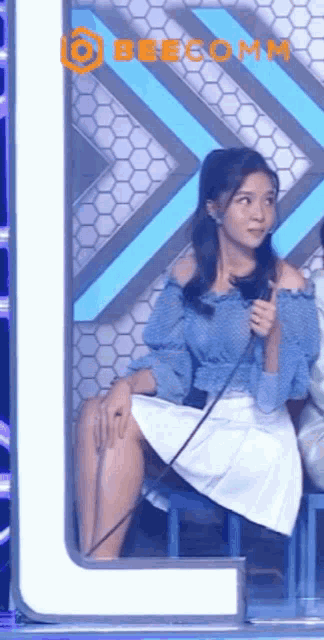 a woman in a blue top and white skirt is sitting on a stool with a microphone in her hand .