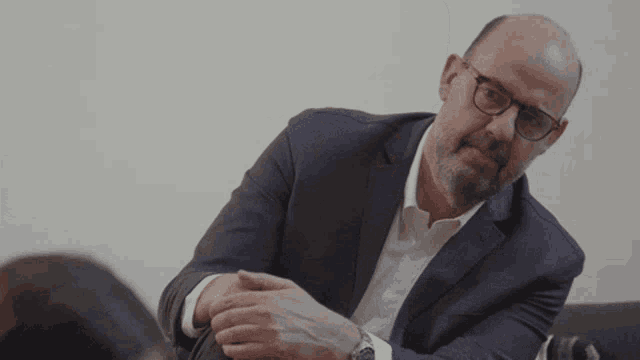 a bald man wearing glasses and a suit looks at the camera