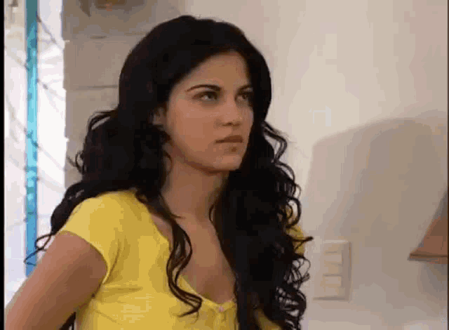 a woman with long black hair and a yellow shirt is standing in front of a window .