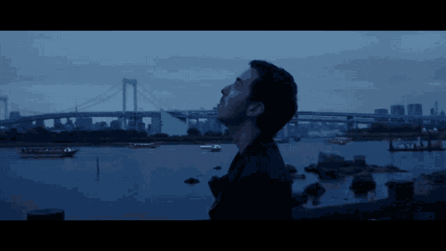 a man stands in front of a large body of water with a bridge in the background