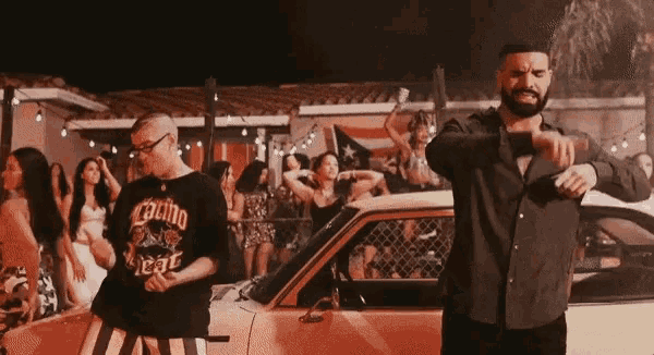 two men are dancing in front of a car and one has a shirt that says calibo