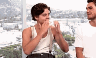 two young men are standing next to each other in front of a window . one of the men is eating a sandwich .