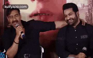 two men are sitting next to each other and one is singing into a microphone while the other is hugging him .