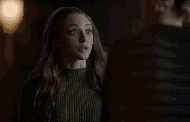 a woman in a green sweater is standing in a dark room and looking at the camera .