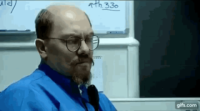 a bald man with glasses and a beard is standing in front of a whiteboard .