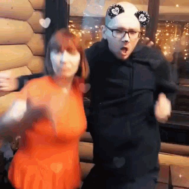 a man and a woman are dancing together in front of a log cabin . the woman is wearing an orange dress .