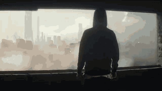 a man in a hoodie is standing on a balcony looking out at a city .