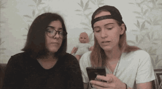 two women are looking at a cell phone together . one of the women is wearing glasses and a baseball cap .